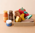 A wooden toy food and drink play set arranged on a light wood tabletop against a warm beige background. The set includes toy bottles labeled Juice, Choc Milk, and Blueberry Lemonade, plus a tray with toy fruit and bread pieces such as a strawberry, banana, apple, avocado, bread, and chocolate bar.